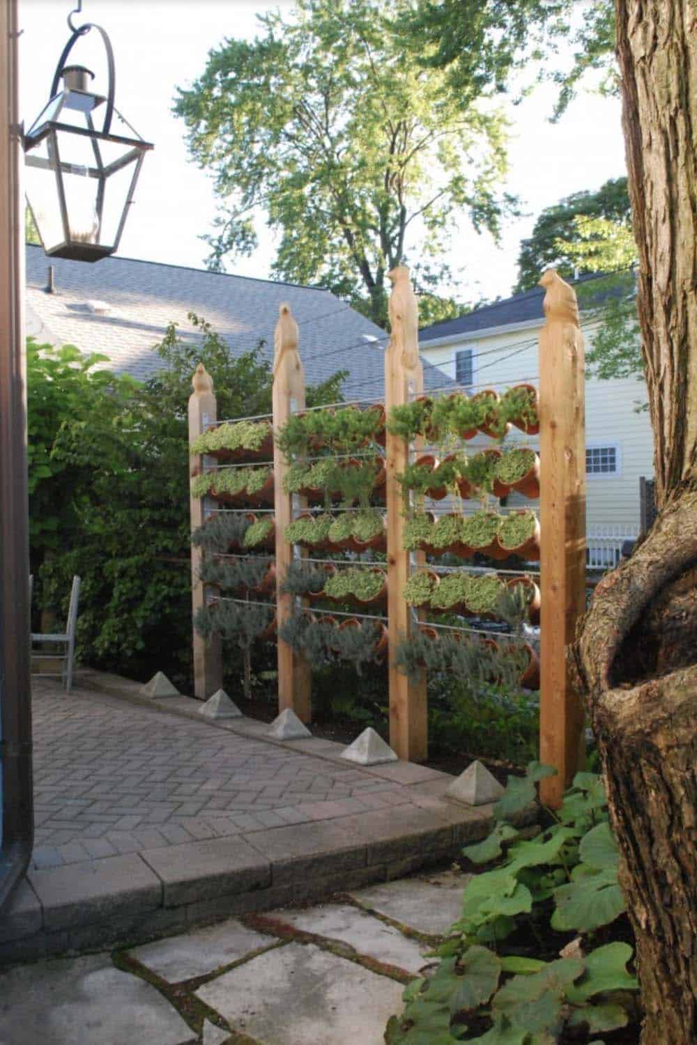 Vertical privacy screen made of cedar posts with tiered wire rows holding terracotta pots of herbs and succulents beside a brick paver patio
