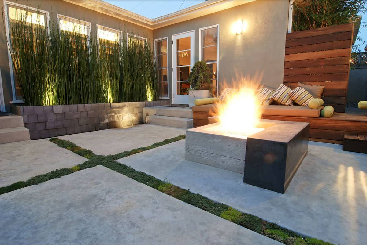 Modern concrete patio with square fire pit, built-in wood bench seating, tall ornamental grasses in raised planters, and a horizontal cedar privacy fence at dusk