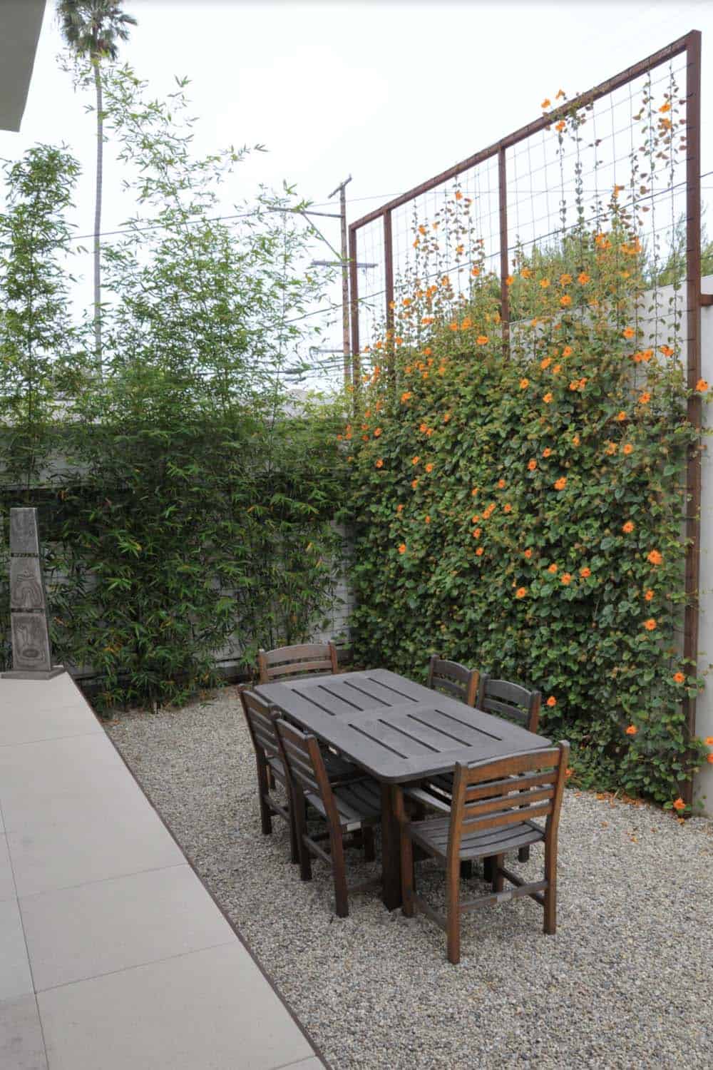 Outdoor dining area on gravel with bamboo and an orange flowering vine climbing a tall metal trellis screen