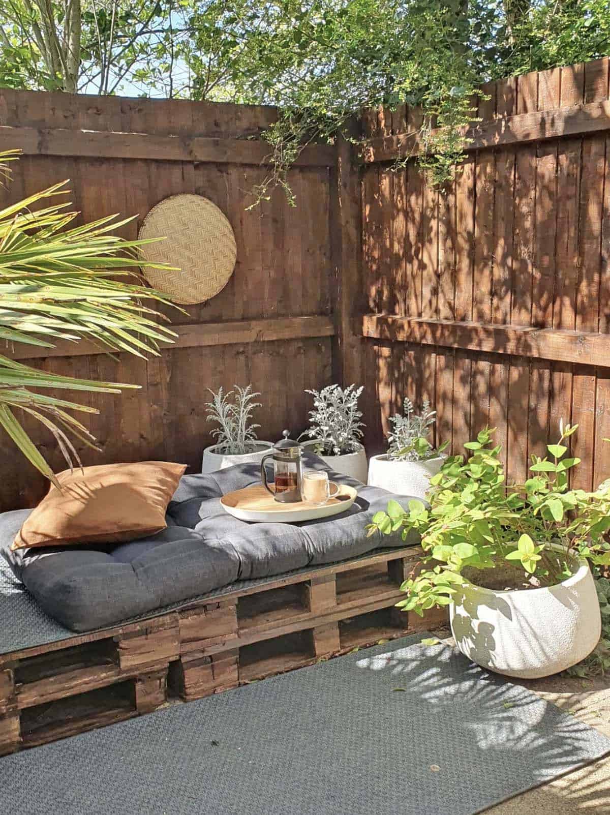 Boho garden nook with stacked wood pallet daybed, charcoal cushions, potted herbs, and a woven wall decoration against a dark stained wood fence