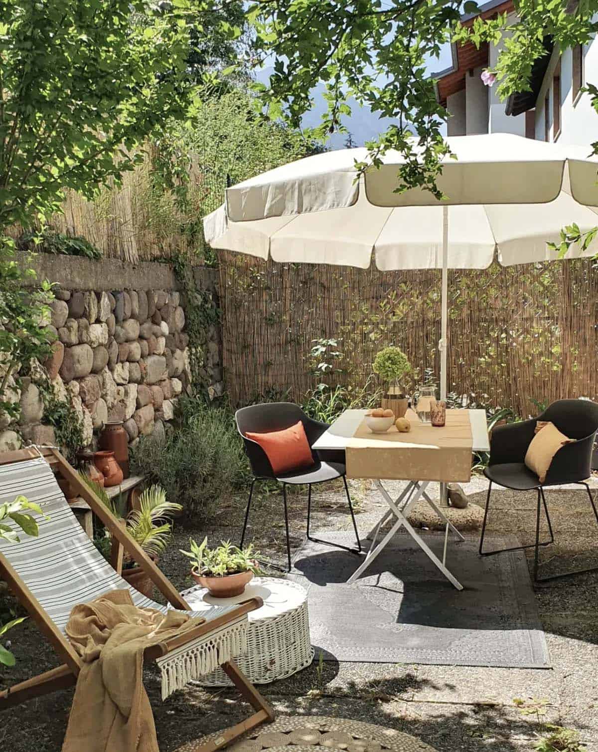 Sun-drenched garden patio with reed roll privacy screen, stone wall, deck chair, and black dining chairs around a white folding table under a cream umbrella