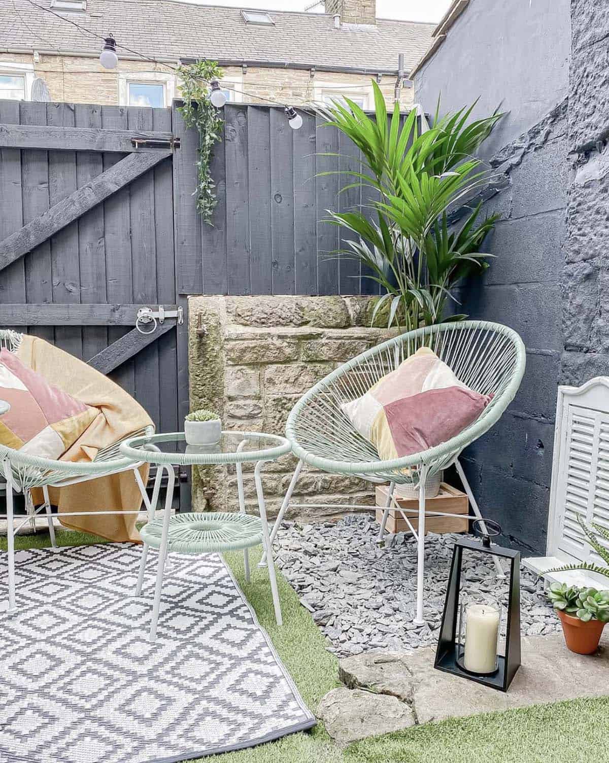 Small urban courtyard in Lancashire with dark painted fence and wall, mint green Acapulco chairs, palm plant, string lights, and a monochrome outdoor rug
