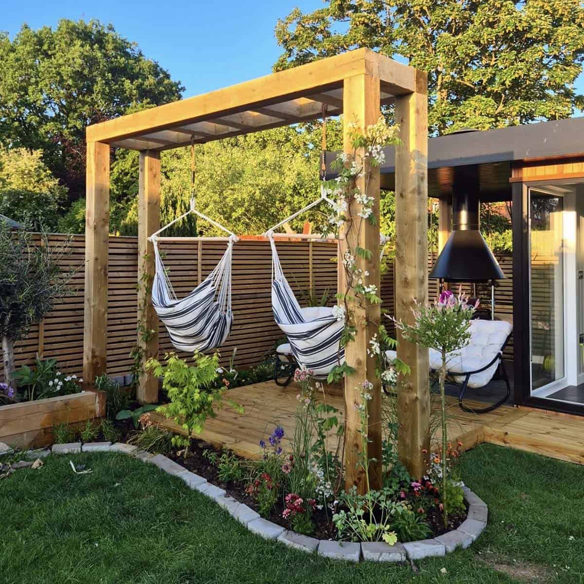 DIY timber pergola with two striped hammock chairs, horizontal slatted wood fence, flowering border, and a modern garden room beyond