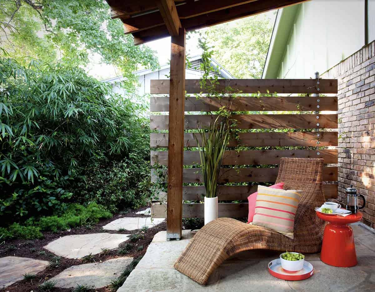 Covered patio nook with horizontal slatted wood screen, wicker chaise lounge, red side table, and bamboo garden beyond