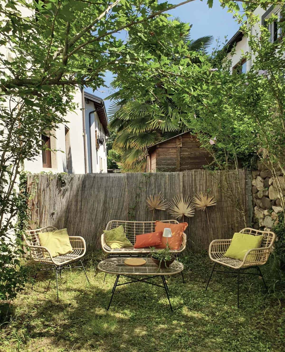 Boho garden seating area with rattan chairs and sofa, reed fence backdrop, dried palm leaf wall decor, and lime green and terracotta cushions