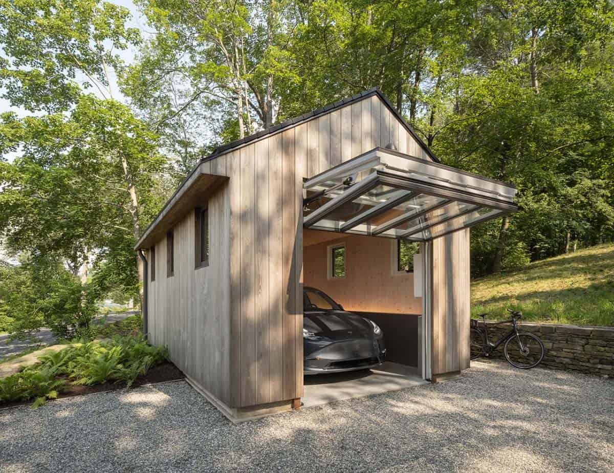 Cedar-clad prefab garage with glass door open at an angle, Tesla inside, and bicycle leaning against a stone wall