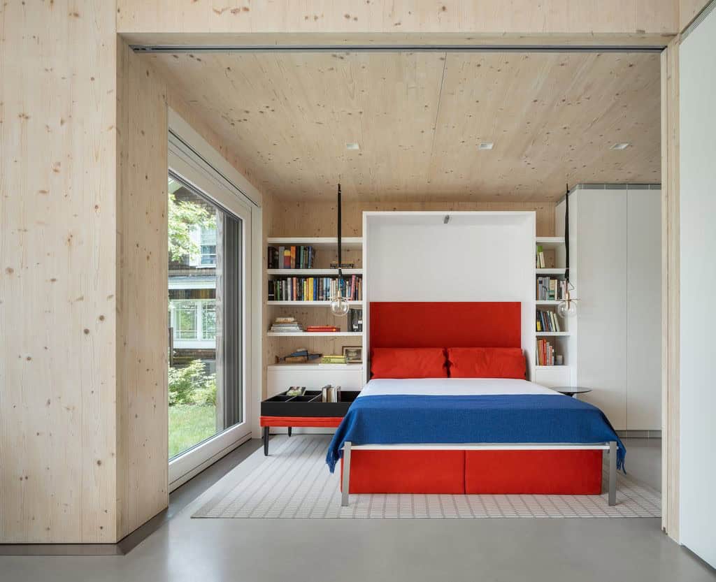 Murphy bed with red headboard and built-in bookshelves in a CLT-paneled bedroom