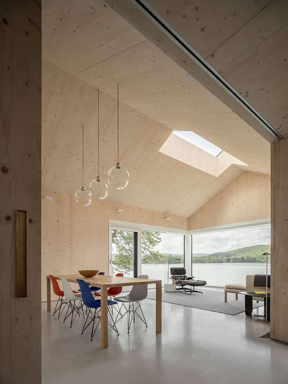 Open-plan dining and living room with CLT ceiling, colorful Eames chairs, skylight, and lake views