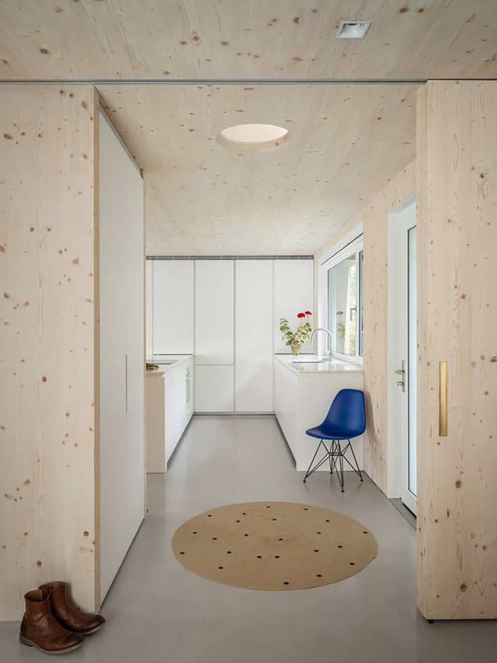 CLT-lined entryway leading to a minimalist white kitchen with a blue Eames chair