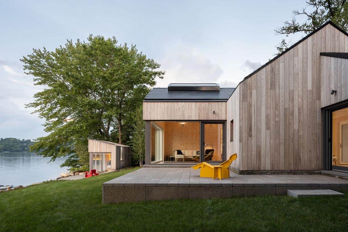 Rear terrace of prefab CLT lake house at dusk with open living area and detached guest studio beyond