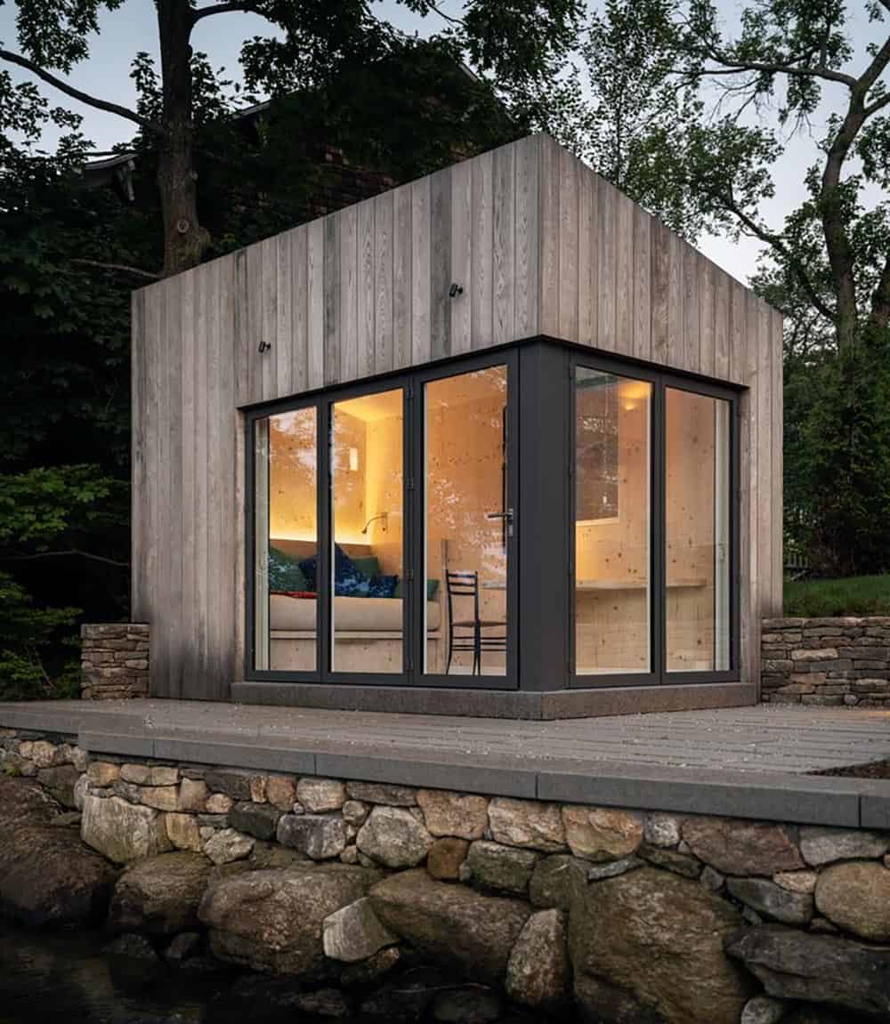 Detached prefab CLT guest studio at dusk with corner glass doors and stone retaining wall