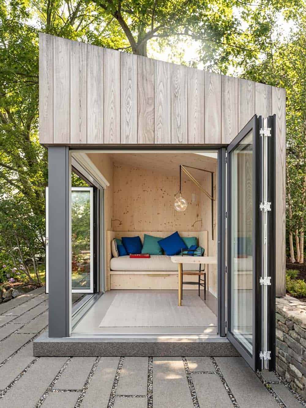 Prefab CLT writer's studio with bifold glass doors open to a stone patio surrounded by trees