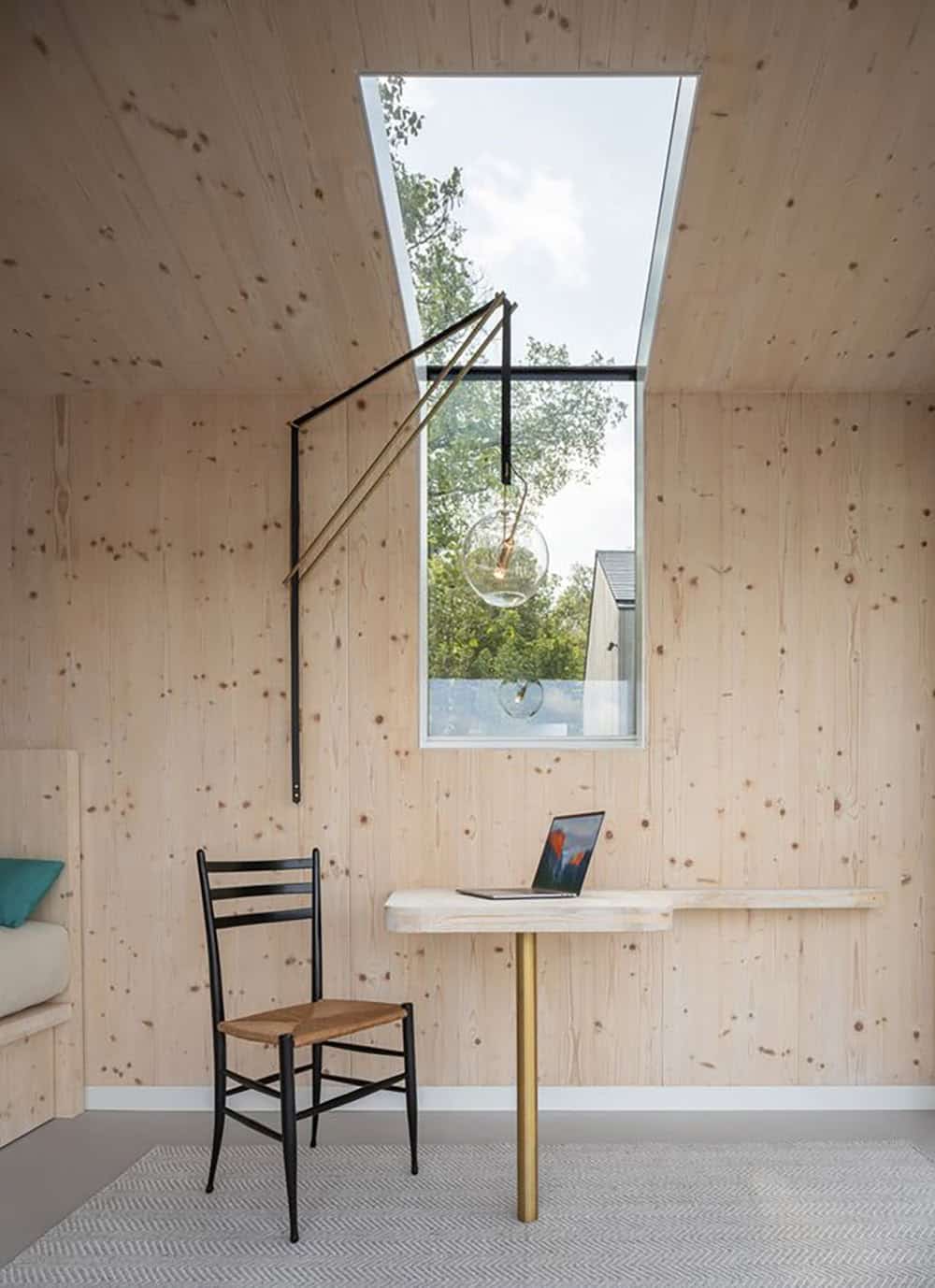 CLT studio workspace with wall-mounted brass pendant light, fold-down desk, and tall skylight window overlooking the main house