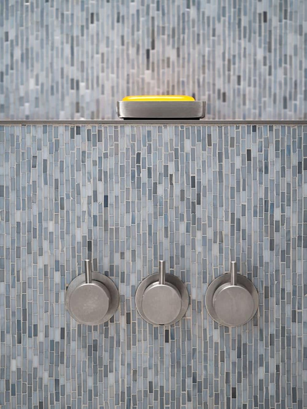 Three stainless shower controls against blue and gray mosaic tile with a yellow soap dish