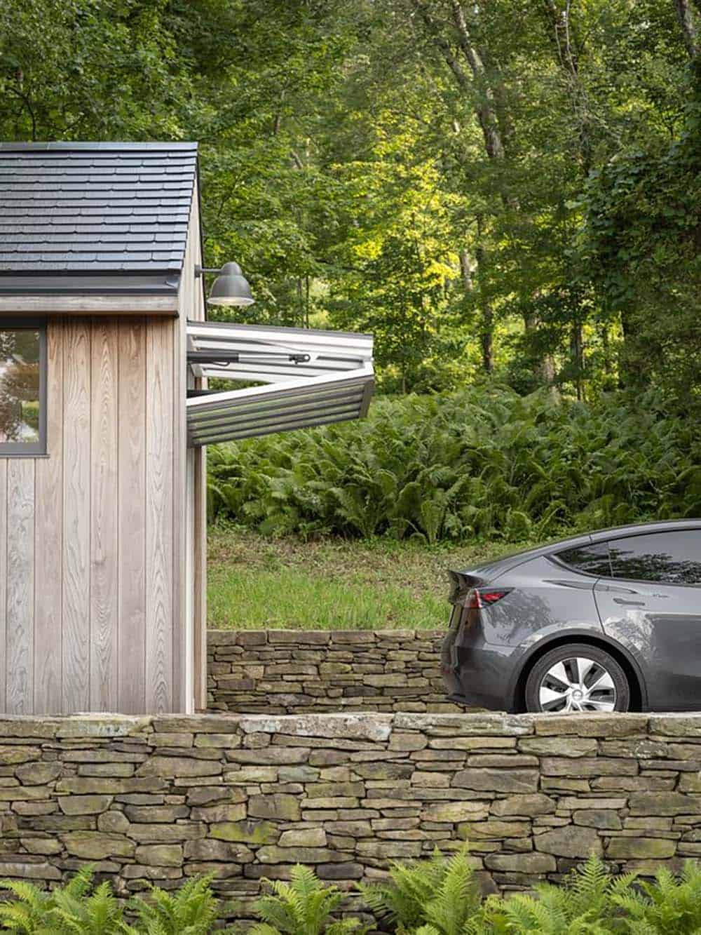 Tesla electric car beside a cedar-clad outbuilding with a stone retaining wall and fern plantings