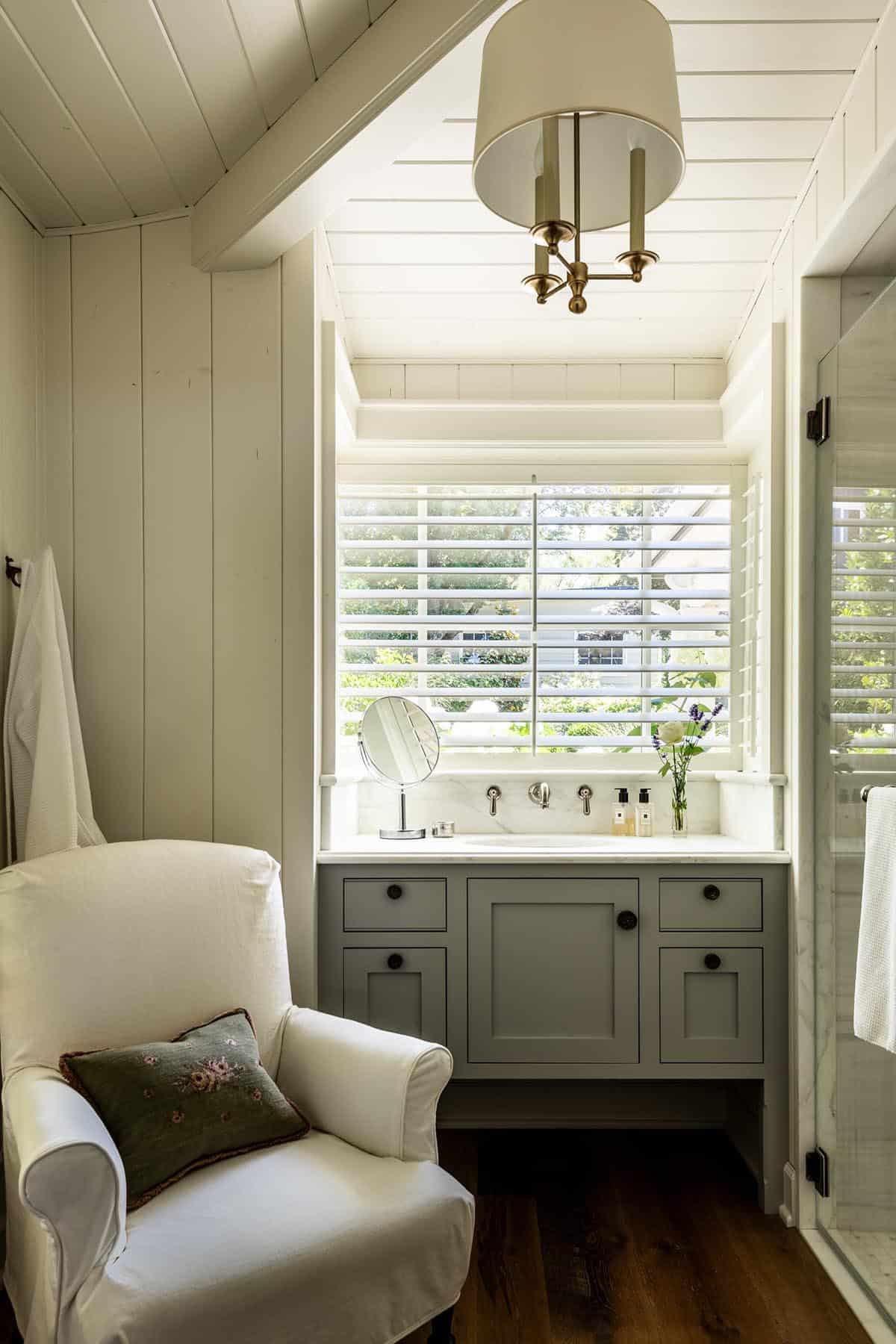 Primary bath with sage vanity, plantation shutters, brass fixture, and white armchair