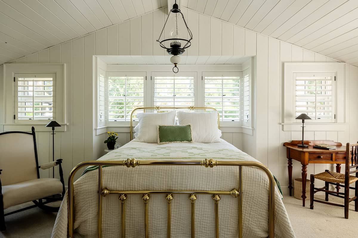 Bedroom with brass bed frame, cathedral ceiling, plantation shutters, and antique desk