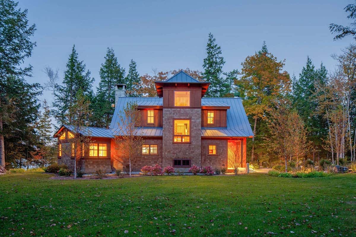 Rear view of three-story cedar shingle camp at twilight with standing seam metal roof and lit interior