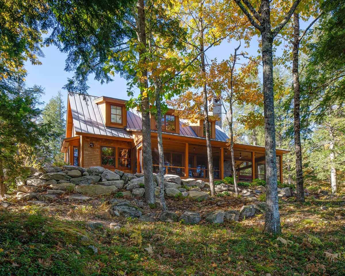 Side view of rustic Maine camp perched on rocky hillside with screened porch and metal roof amid fall foliage