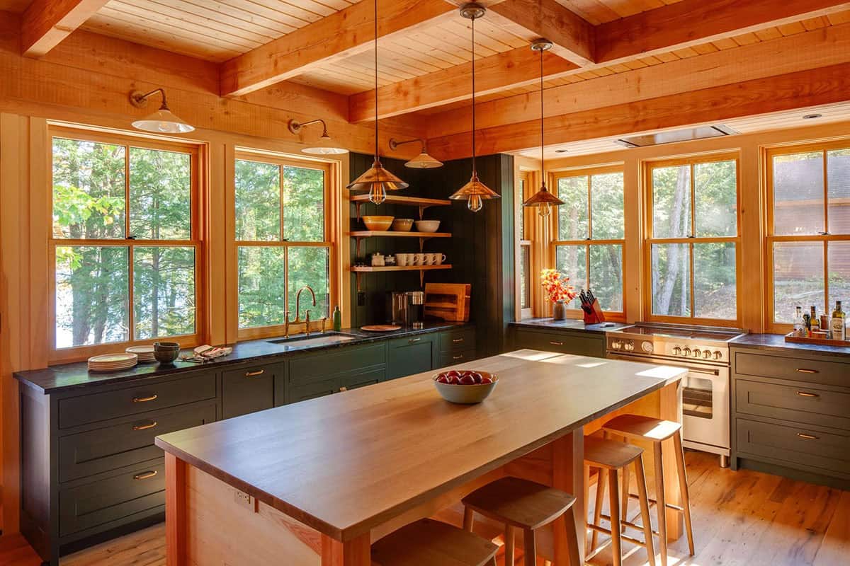 Rustic kitchen with dark green cabinets, wood island, exposed beam ceiling, copper pendant lights, and forest views