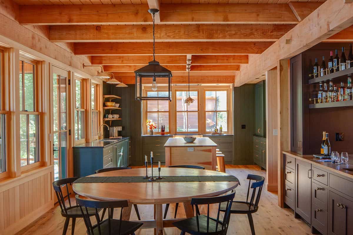 Open-plan dining and kitchen area with round wood table, dark Windsor chairs, exposed beams, and bar nook