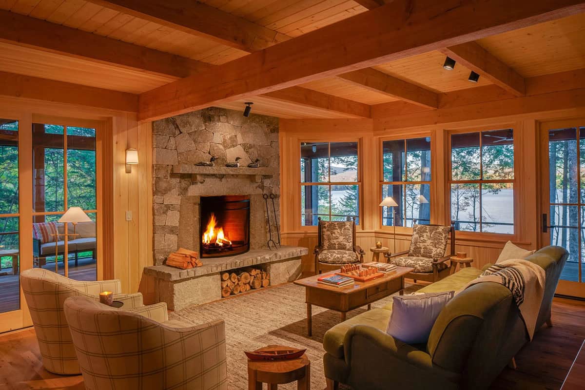 Living room with granite fireplace, exposed timber beams, mixed seating, and large windows overlooking a lake