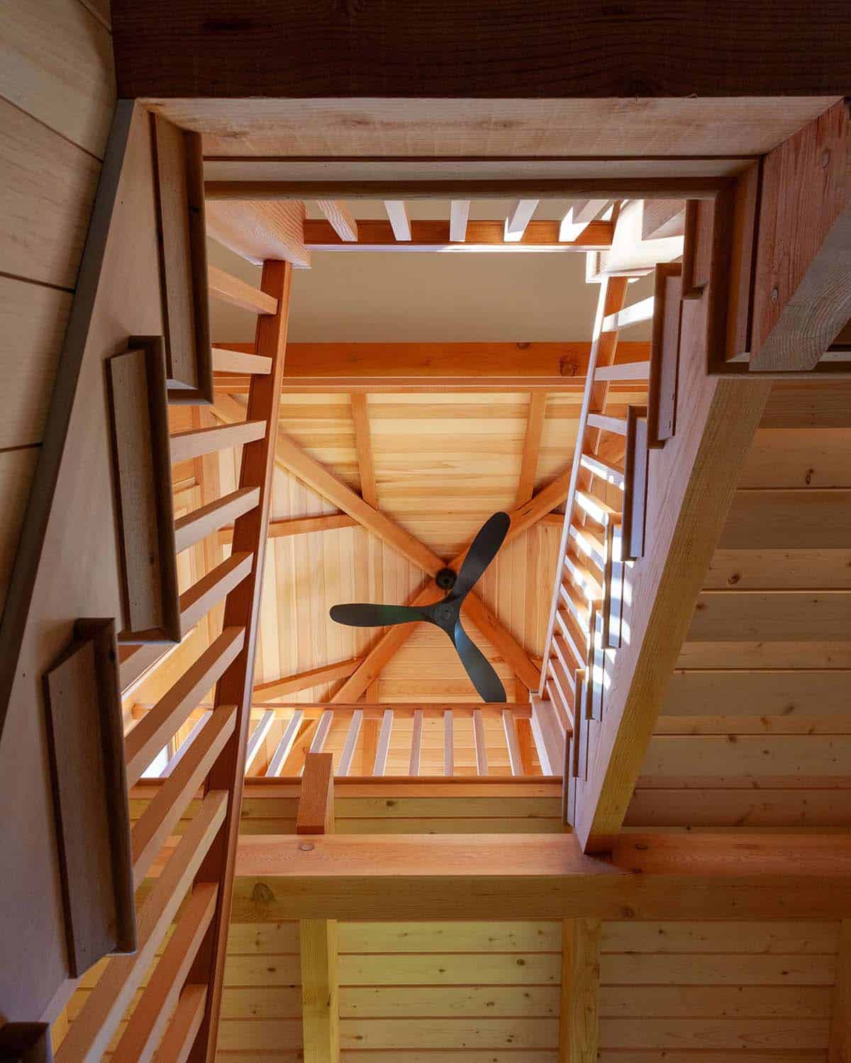 Looking up through stair tower well showing spiraling wood stairs, timber trusses, and ceiling fan