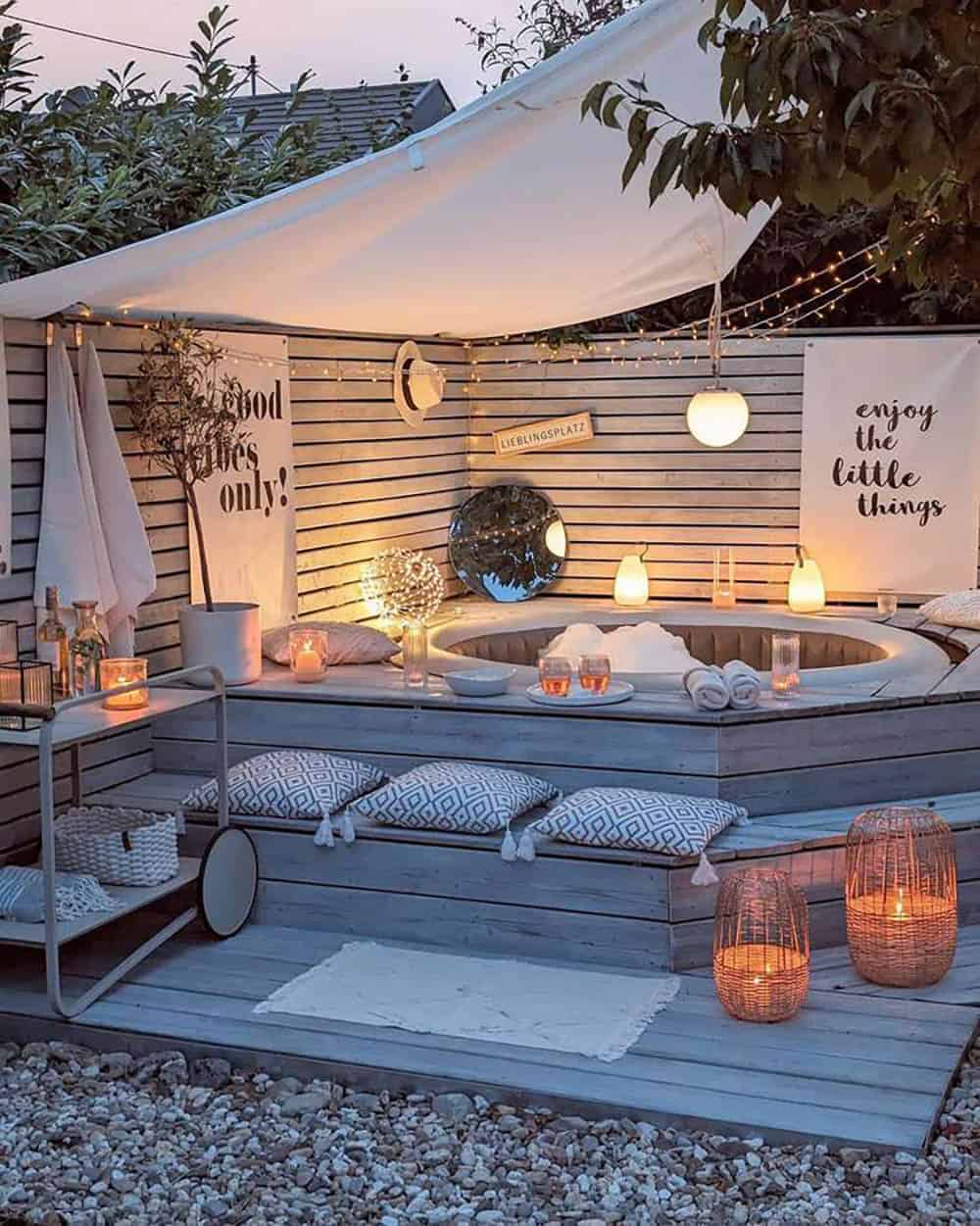 A boho-style hot tub deck at dusk with a canvas canopy, string lights, lanterns, and patterned floor cushions