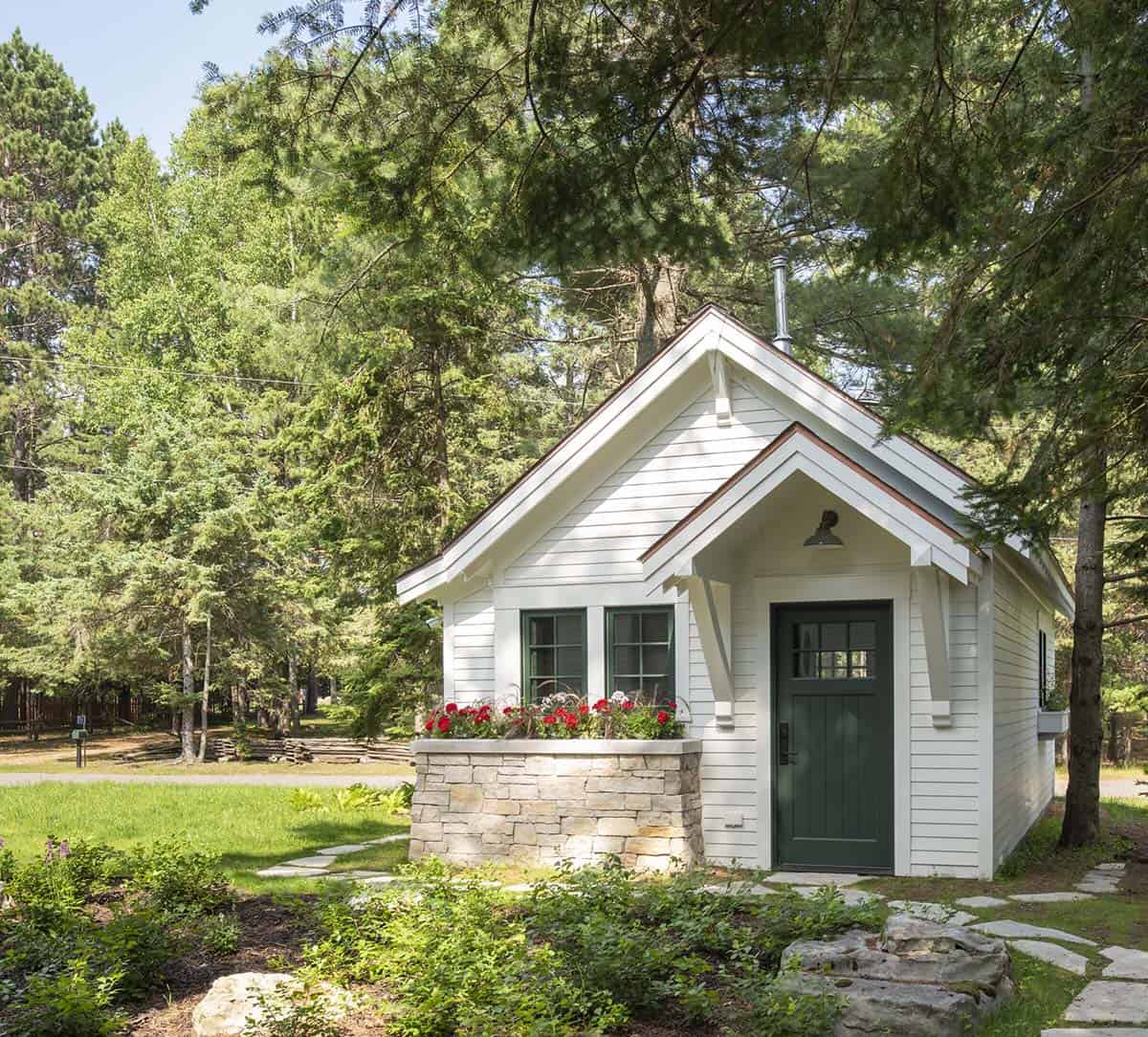 Small white cottage with green door, stone base, and red flower window box surrounded by pine trees