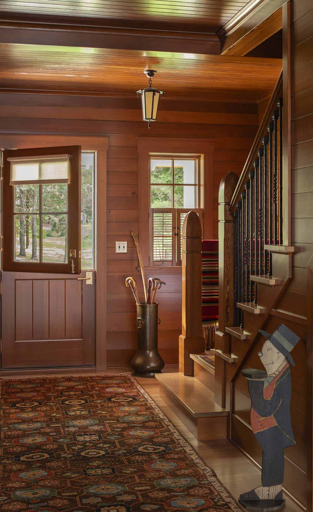 Warm wood-paneled entryway with Dutch door, staircase, hanging lantern, and patterned area rug