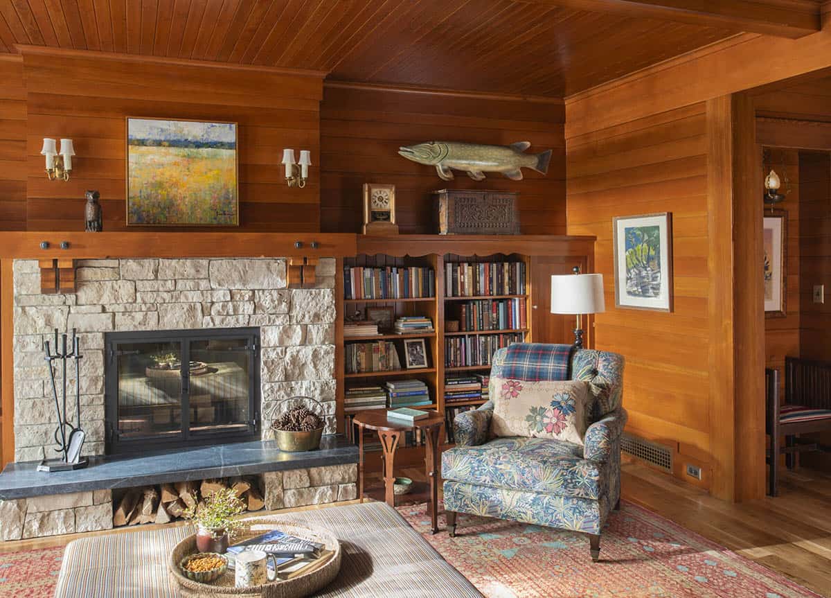 Living room with stone fireplace, built-in bookshelves, floral armchair, and cedar-paneled walls