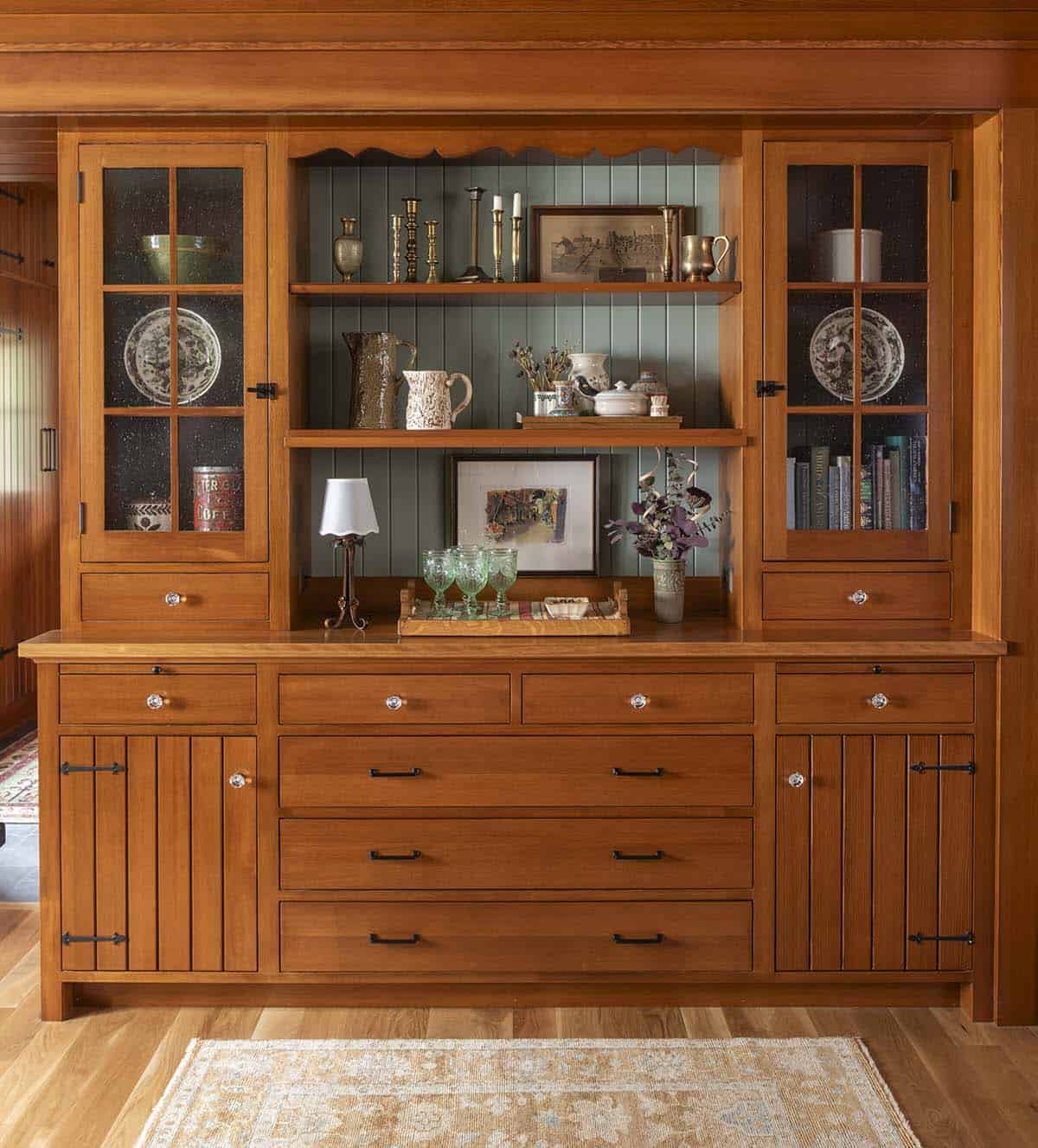 Built-in wood hutch with glass cabinet doors, open shelving, and sage green painted back panel