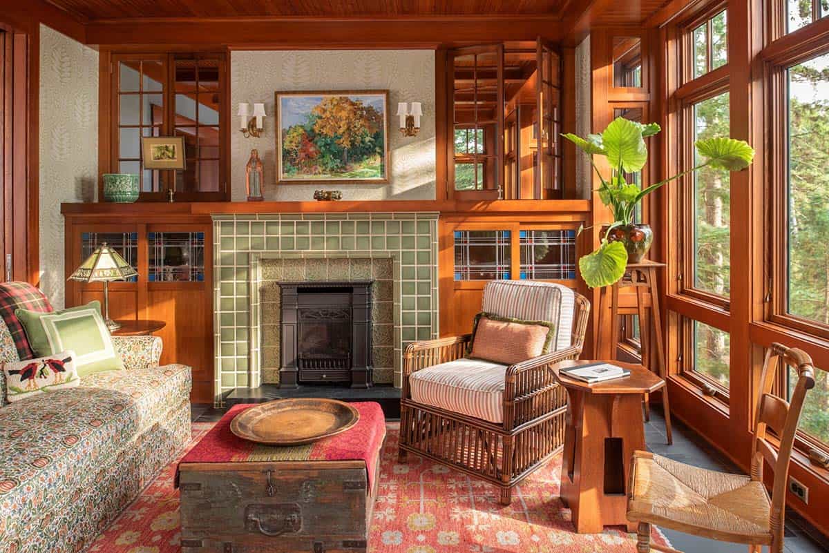 Sun-filled sitting room with green tile fireplace, wicker chair, stained glass windows, and forest views