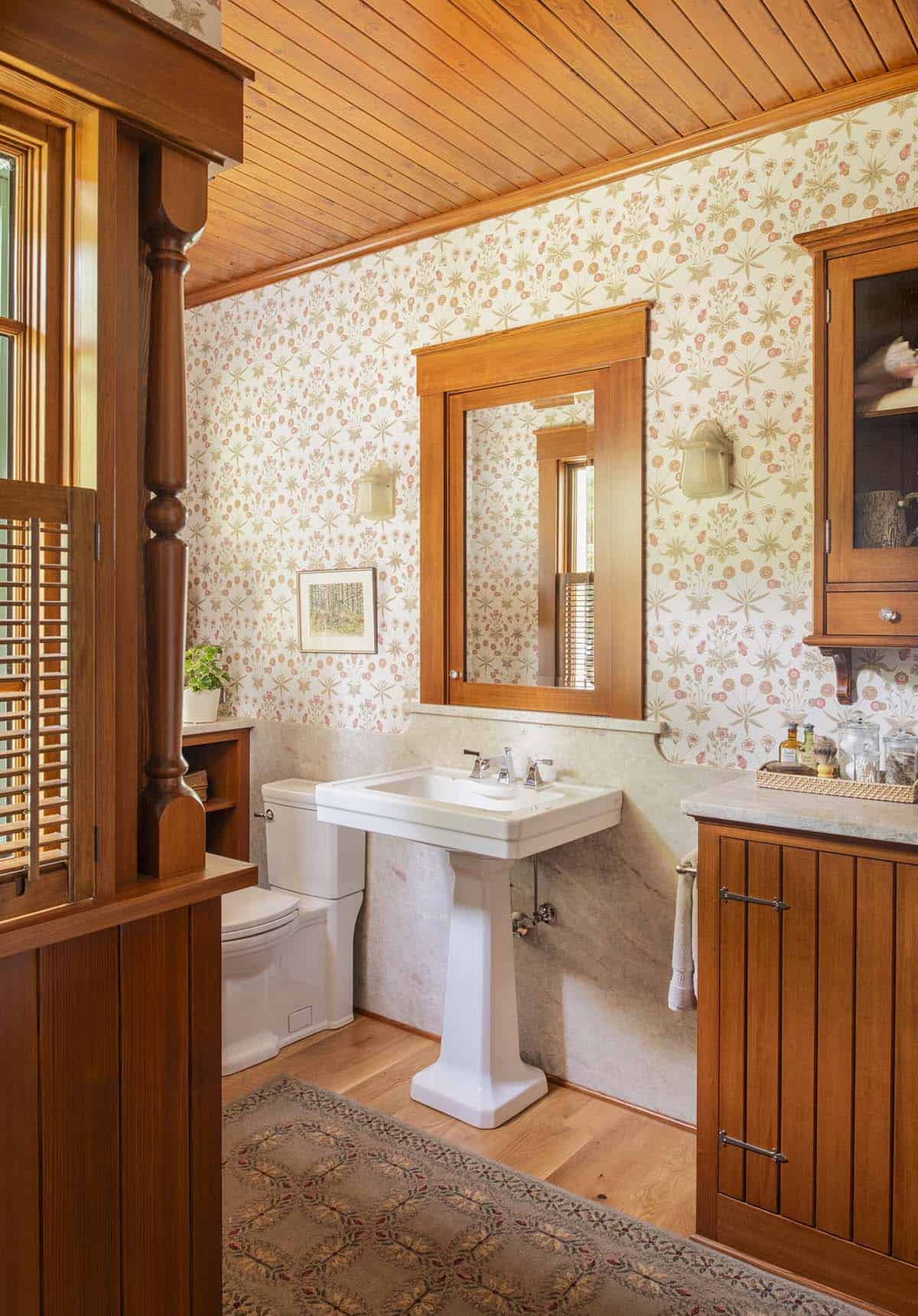Bathroom with pedestal sink, marble wainscoting, floral wallpaper, cedar ceiling, and wood-framed mirror