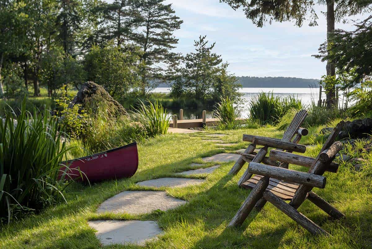 Stone path leading to a wooden dock on a calm lake, with rustic log chairs and a red canoe on shore