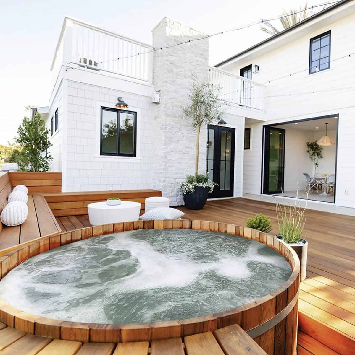 A round cedar hot tub set into a backyard deck that flows seamlessly into a white modern farmhouse with an indoor-outdoor living space