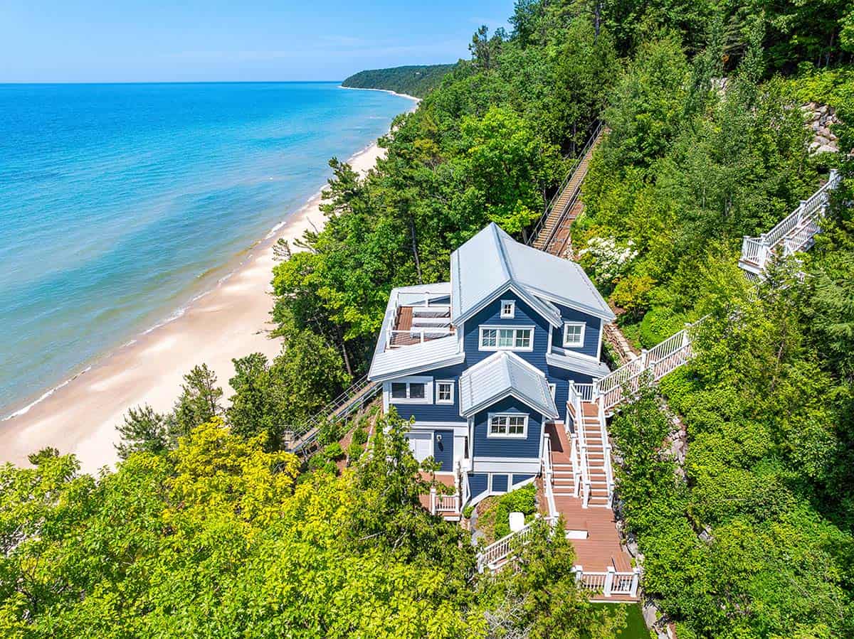 Aerial view of blue shingle-style house on a wooded bluff overlooking Lake Michigan's turquoise water and sandy beach