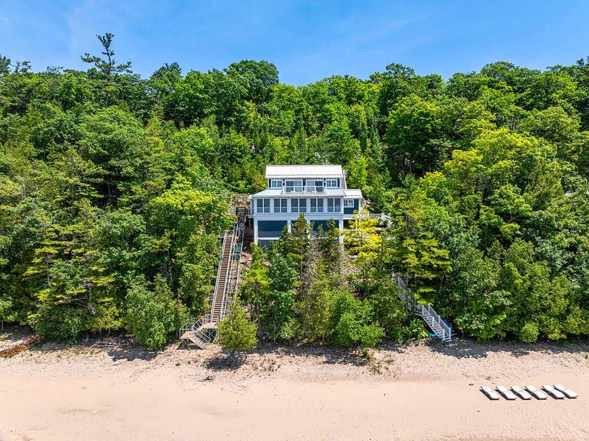 Aerial view of a white cottage with metal roof nestled in trees above a sandy Lake Michigan beach with lounge chairs