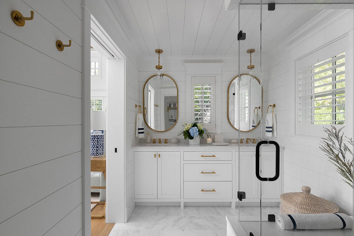 Bright white bathroom with double vanity, oval brass mirrors, pendant lighting, and glass shower enclosure