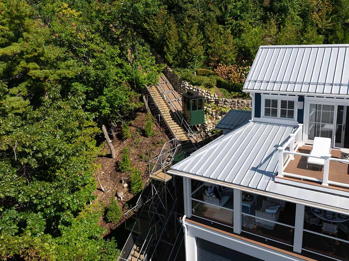 Aerial view of a funicular tram on tracks running through the wooded cliffside beside the house's metal roof and deck