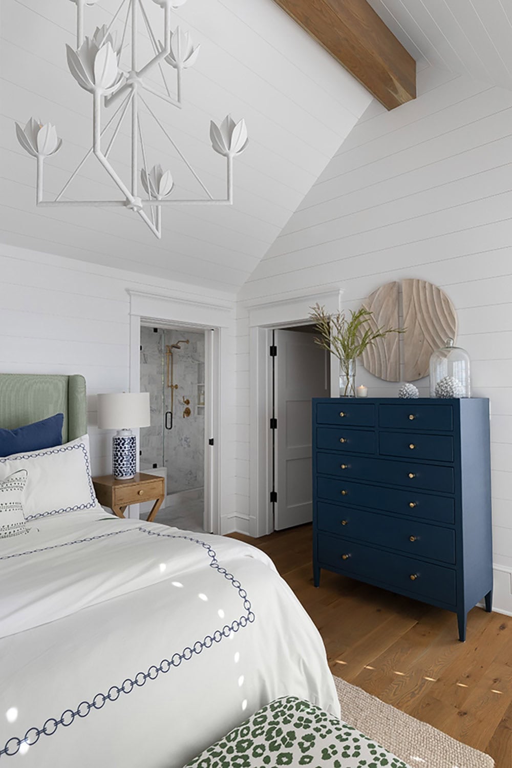 Bedroom with vaulted shiplap ceiling, exposed wood beam, navy chest of drawers, and view into an en-suite bathroom