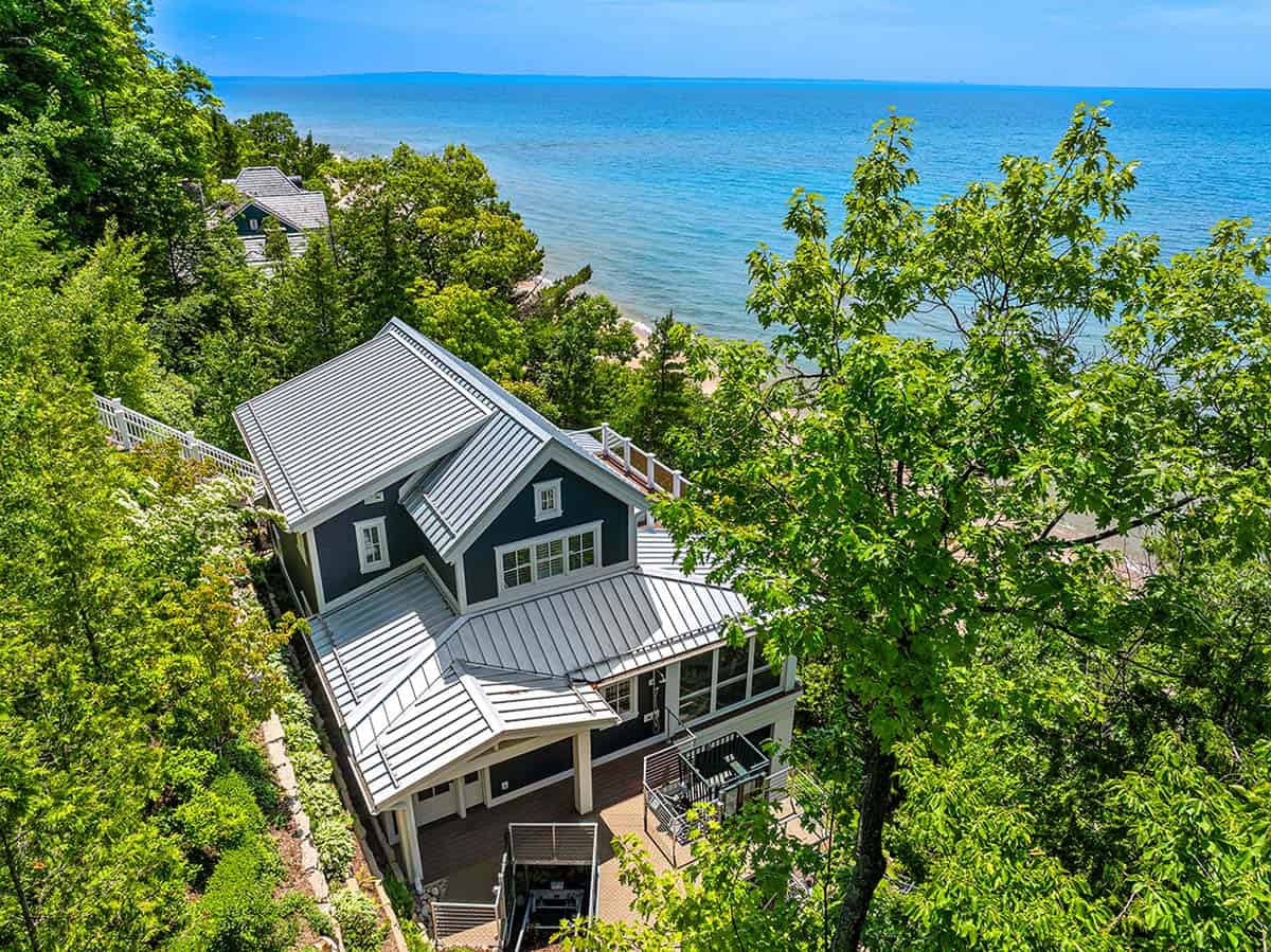 Aerial view of blue cottage with metal gabled roofs perched on a wooded bluff above Lake Michigan's beach
