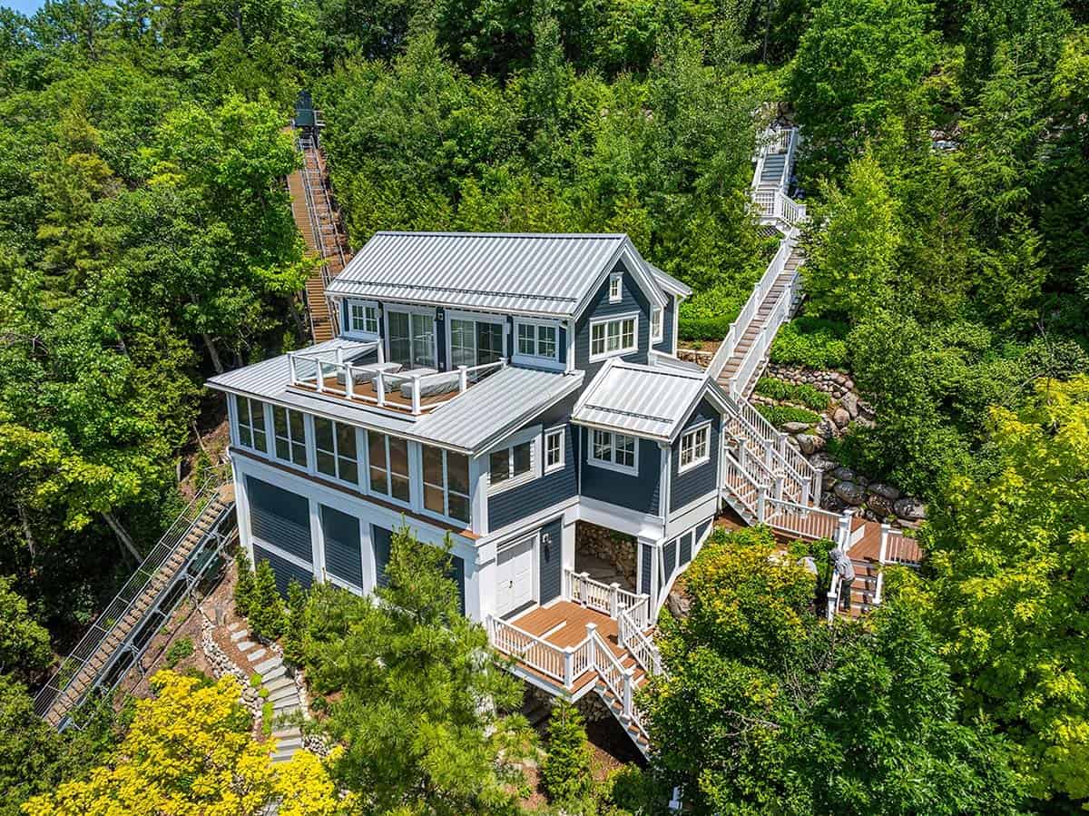 Aerial view of blue cottage with metal roofs and rooftop deck, flanked by long staircases through the forest