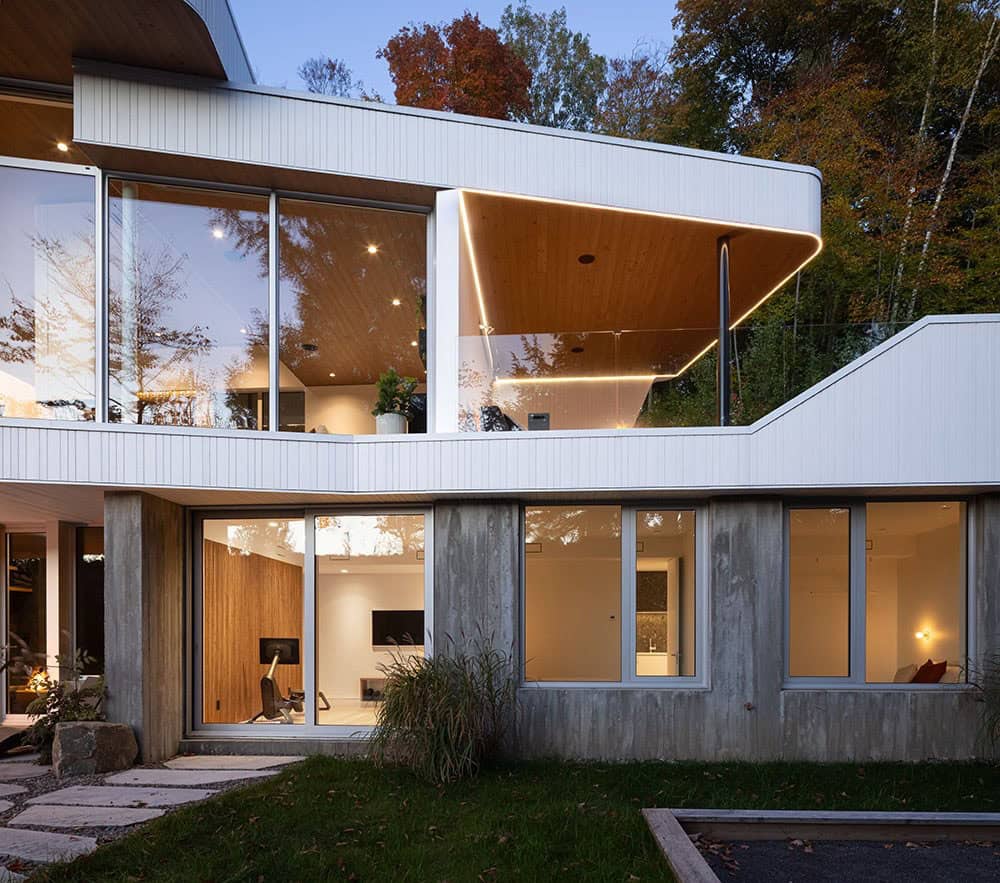 Illuminated exterior at twilight showing cantilevered wood-ceiling overhang and lower-level concrete base with interior views