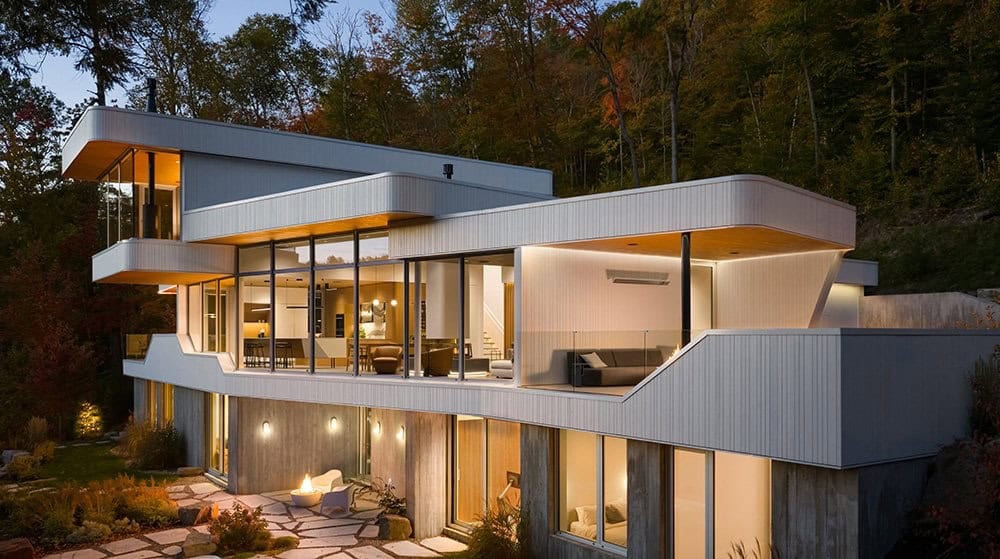 Warm evening view of three-story contemporary home with glowing interiors and stone patio fire feature