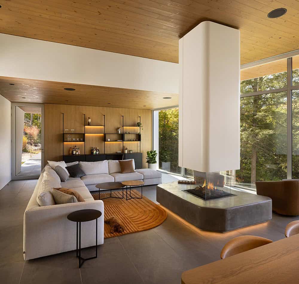 Open-plan living room with three-sided fireplace, sectional sofa, wood ceiling, and floor-to-ceiling forest views