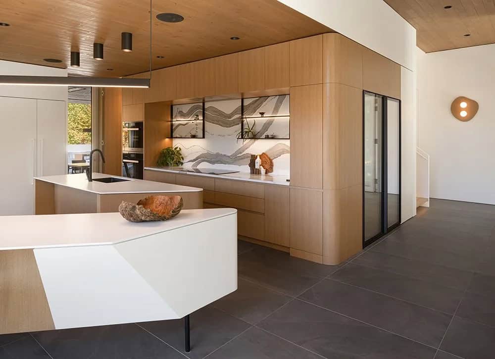 Kitchen overview with sculptural two-tone island, floor-to-ceiling wood cabinetry, and artistic stone backsplash