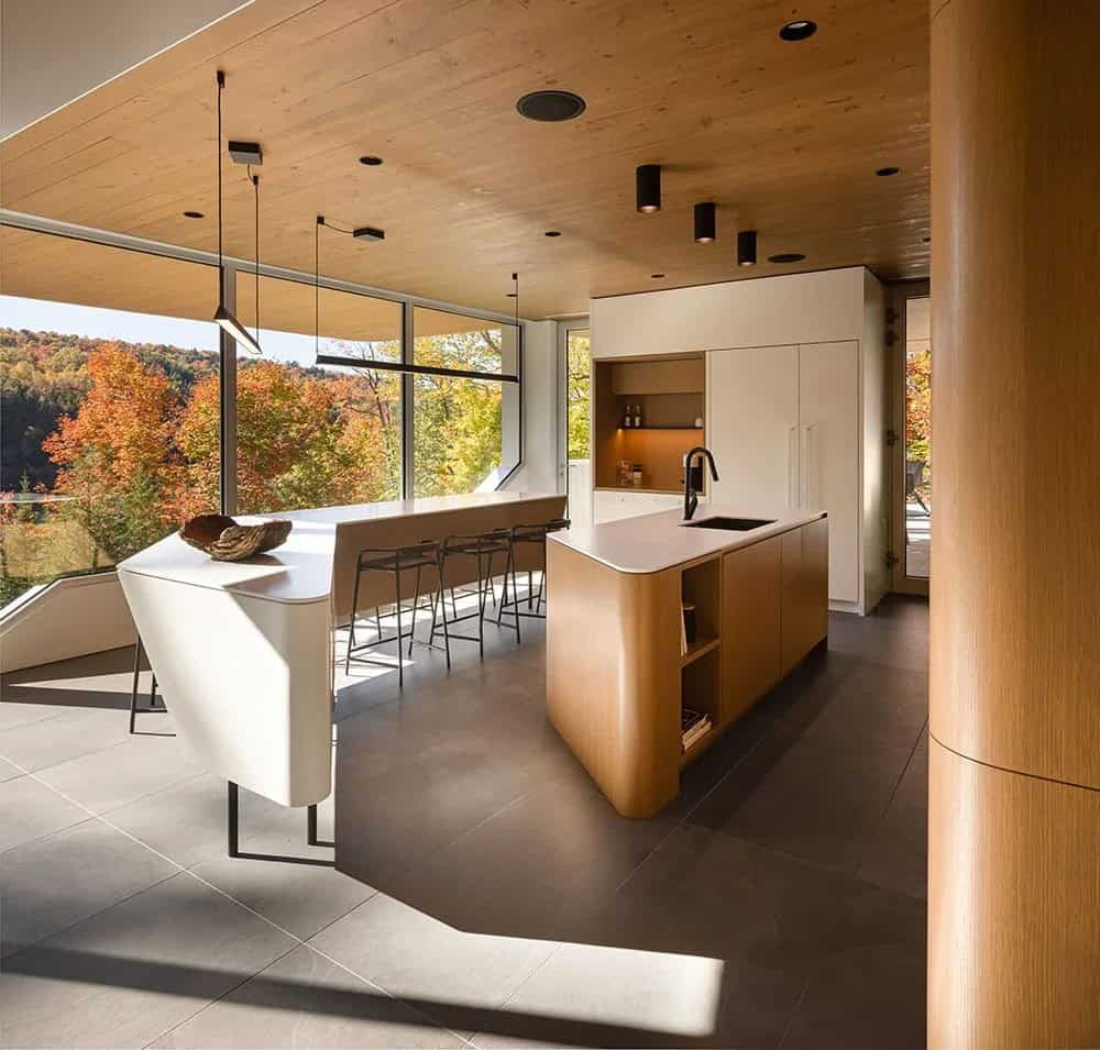 Kitchen with curved wood island and bar seating facing panoramic floor-to-ceiling autumn foliage views