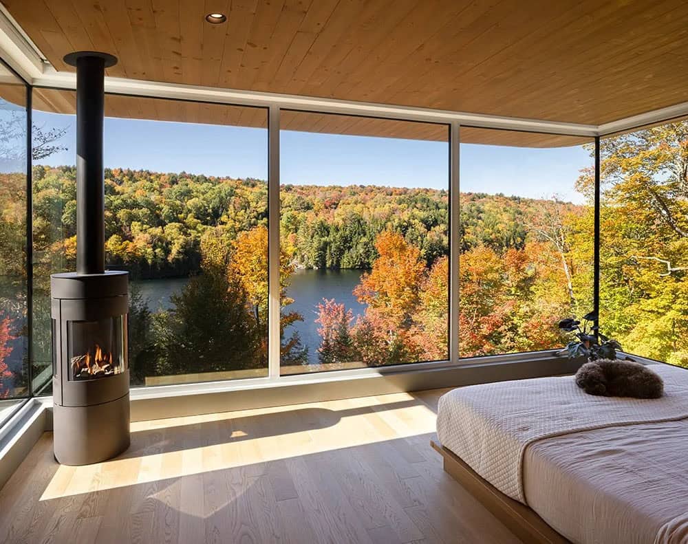 Primary bedroom with freestanding cylindrical wood stove and wraparound windows overlooking lake and fall forest