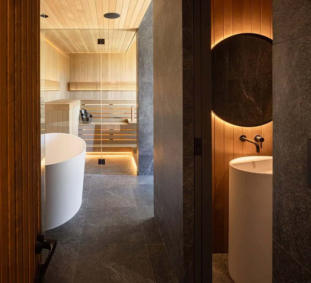 Spa-like lower level bathroom with freestanding tub, round backlit mirror, pedestal sink, and cedar sauna beyond